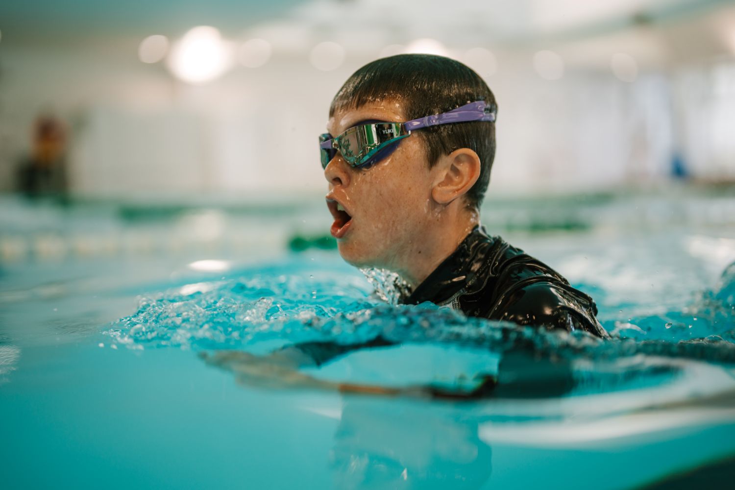 A close-up shows a young boy wearing swim goggles and a rash vest as he surfaces to take a breath while swimming in an indoor pool lane.